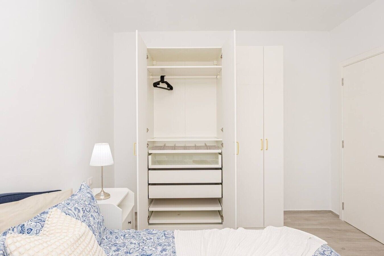 Un dormitorio con cama, una mesita de noche con lámpara, un armario con varios estantes y una puerta.