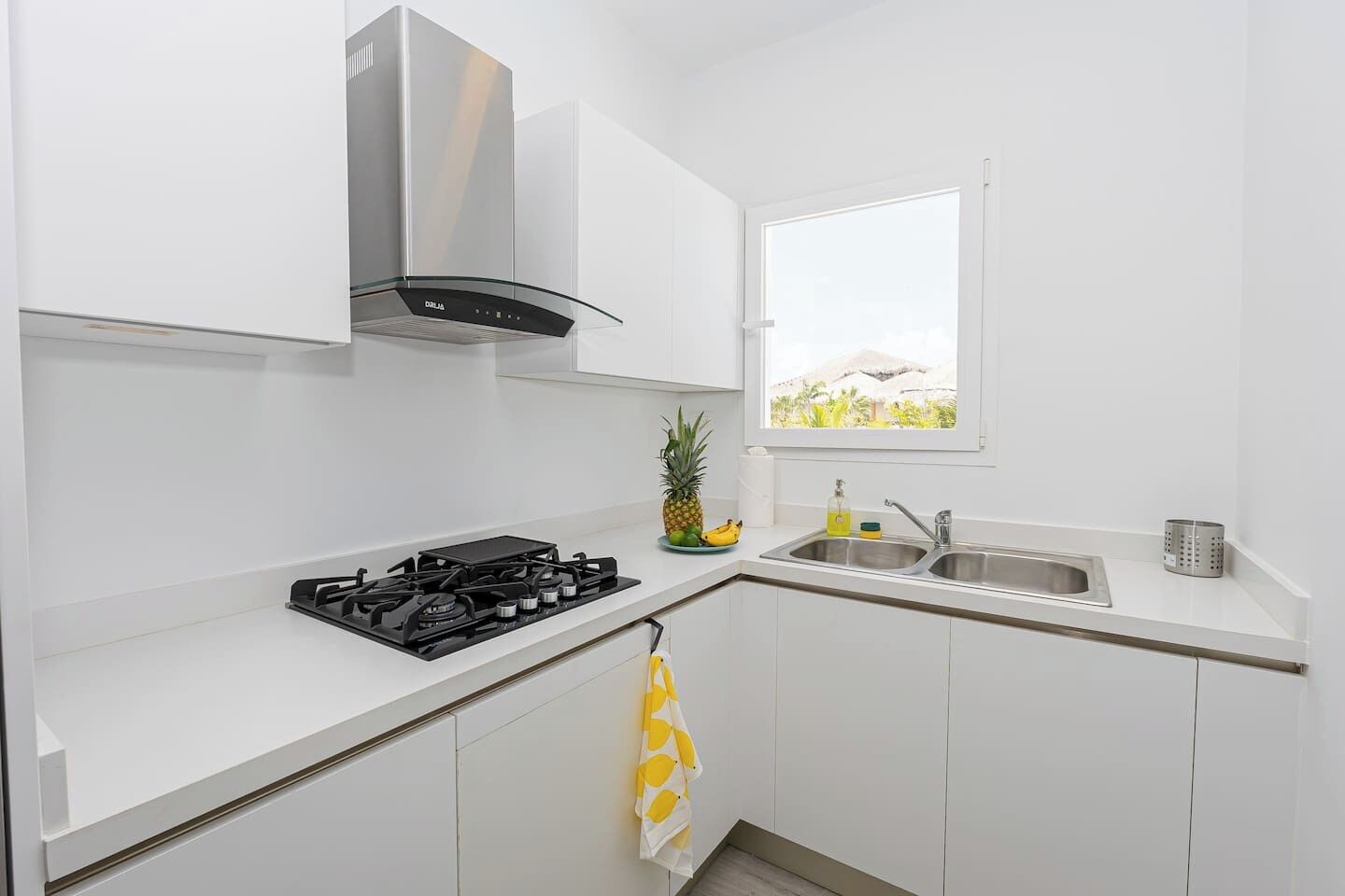 Cocina compacta con armarios blancos, estufa a gas, pileta de acero inoxidable y ventana con vista a vegetación.