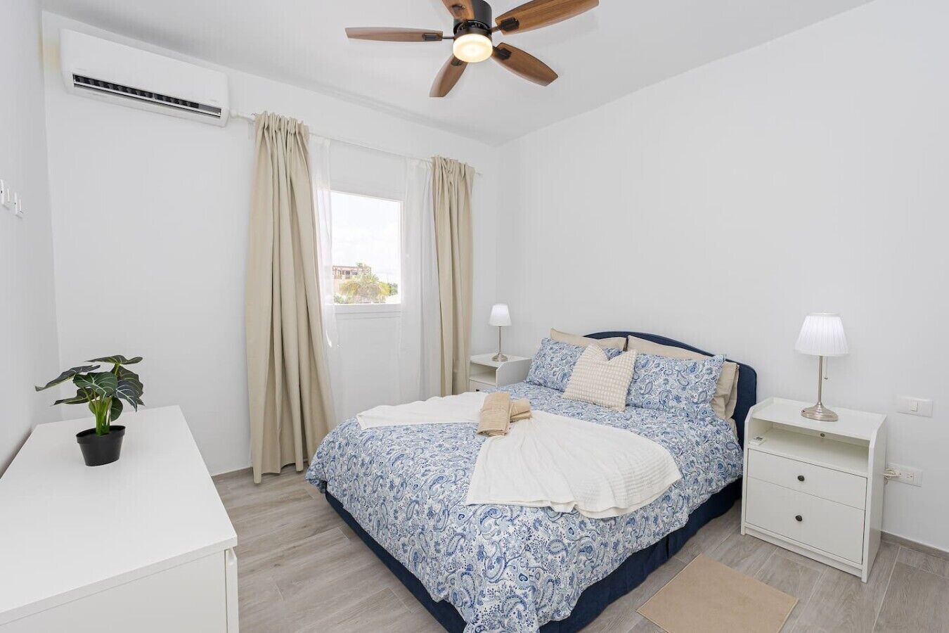 Un dormitorio con una cama, mesitas de noche, ventilador de techo, equipo de aire acondicionado, ventana con cortinas y una planta en maceta.