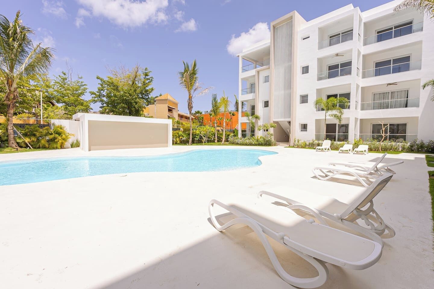 A modern building with a pool, white lounge chairs, and palm trees.