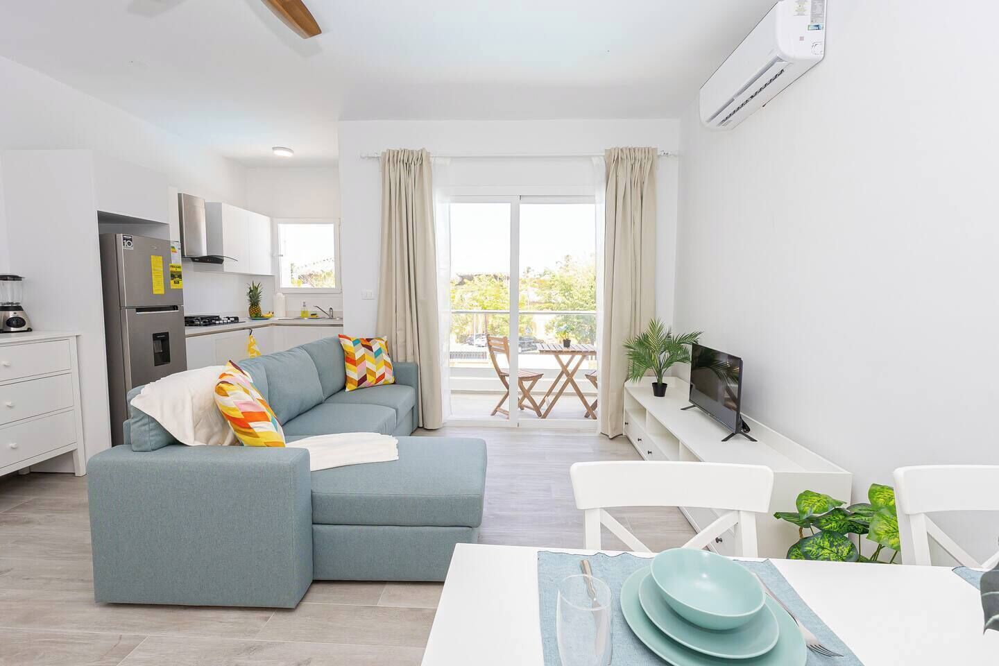 A modern living room with a sofa, a dining table with plates, a TV, and a kitchen area with a refrigerator and stove.