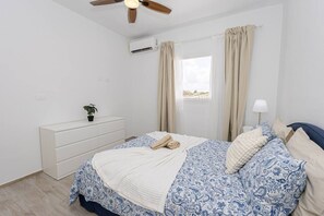 Een slaapkamer met een bed, plafondventilator, airconditioning, raam met gordijnen, bedlamp en een dressoir.