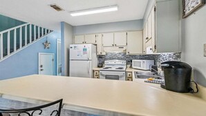 Keittiössä on valkoinen jääkaappi, liesi, mikroaaltouuni ja kahvinkeitin, joka on sijoitettu työtasolle.