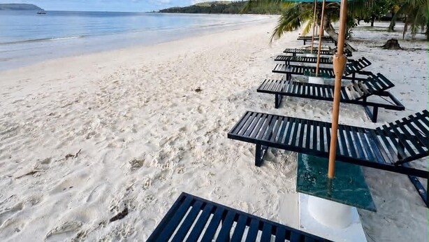 A beach with white sand, a clear blue sky, and a calm sea. There are rows of lounge chairs and umbrellas along the shore. - ARIMATABE Beachfront Resort (Koh Rong)