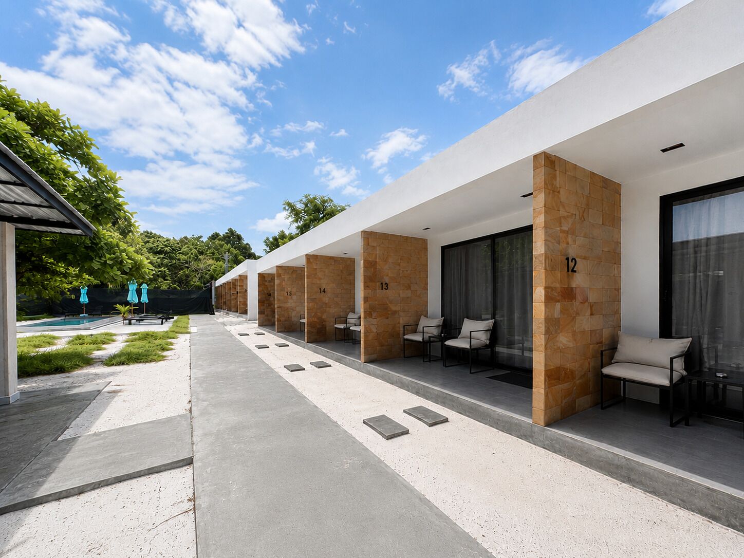 Une rangée de chambres d’hôtel, chacune dotée d’un coin salon, numérotées de 12 à 15, et une piscine en arrière-plan.