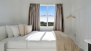 Un dormitorio con cama, almohadas, una mesita de noche, una lámpara colgante y una ventana con cortinas.