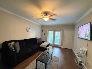 Ein Wohnzimmer mit einem schwarzen Sofa, einem Couchtisch, einem Deckenventilator und einem an der Wand befestigten Fernseher.