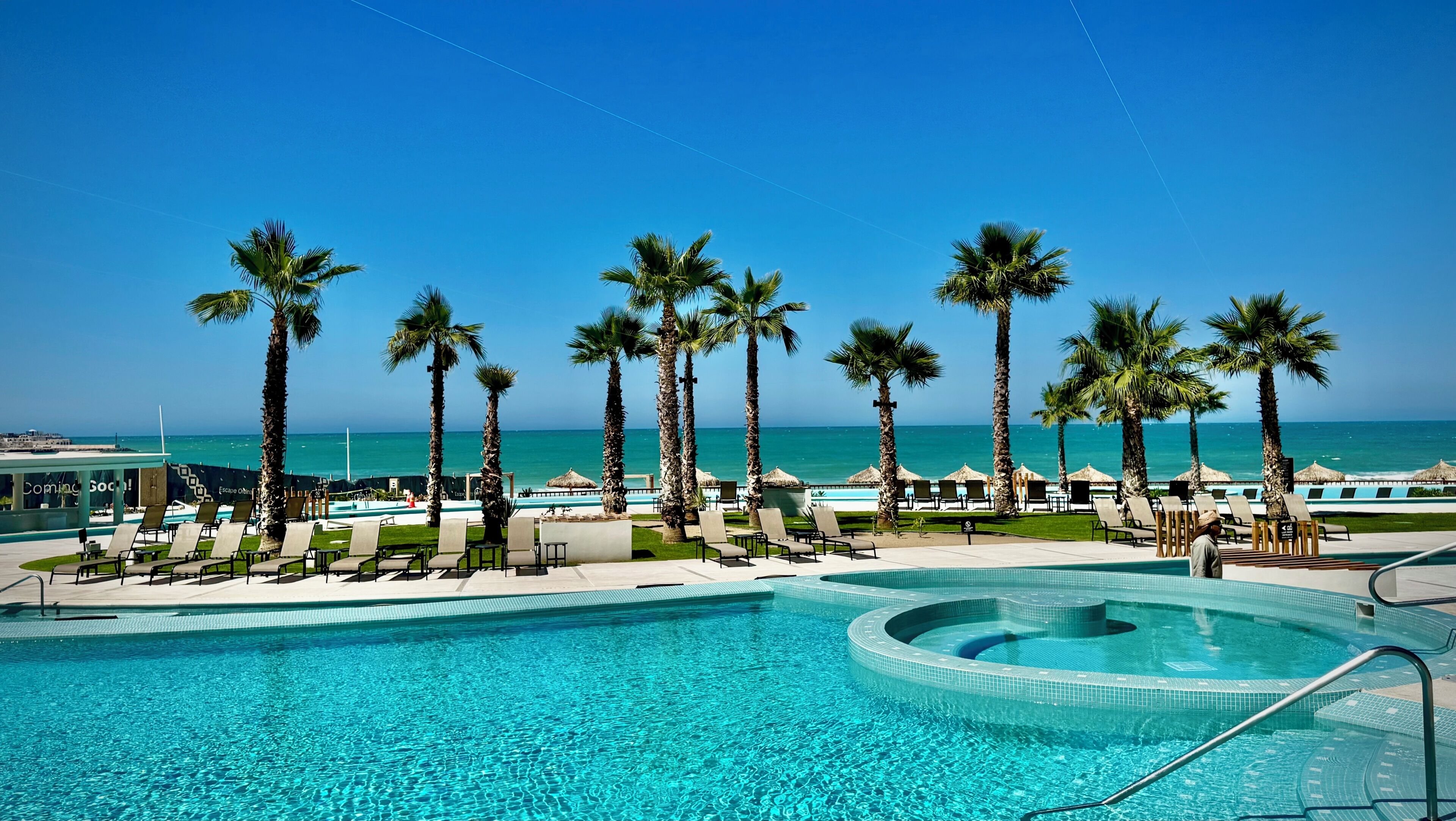A pool area with lounge chairs and palm trees, overlooking a clear blue ocean.