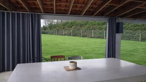 Een overdekt terras met een tafel en stoelen, een uitzicht op een groen gazon en een houten balkenplafond.