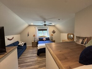 Ein Schlafzimmer mit einem Bett, einem Schreibtisch, einem Stuhl, einem Deckenventilator, einem Fenster mit Vorhängen, einem an der Wand montierten Fernseher und einem Bücherregal.
