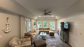 A living room with a sectional sofa, a patterned armchair, a coffee table, a television, and a ceiling fan. - Premium Waterfront 5BR/3.5BA. Boat House, Kayaks, Lillypad, Wheelchair-friendly (Littleton)