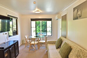 A room with a dining table, chairs, a sofa, a television, and a window with a view of trees. - Sunshine Retreat - Sky Suite (Bli Bli)