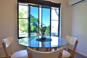 A round glass table with two white chairs, a vase with flowers, and a view of trees through a window. - Sunshine Retreat - Sky Suite (Bli Bli)