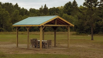 A wooden pavilion with a metal roof, surrounded by chairs and a table, set in a grassy field with trees in the background.
