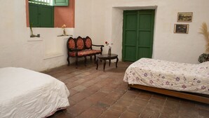 A room with two beds, a chair, a small table, a green door, and a window with green shutters.