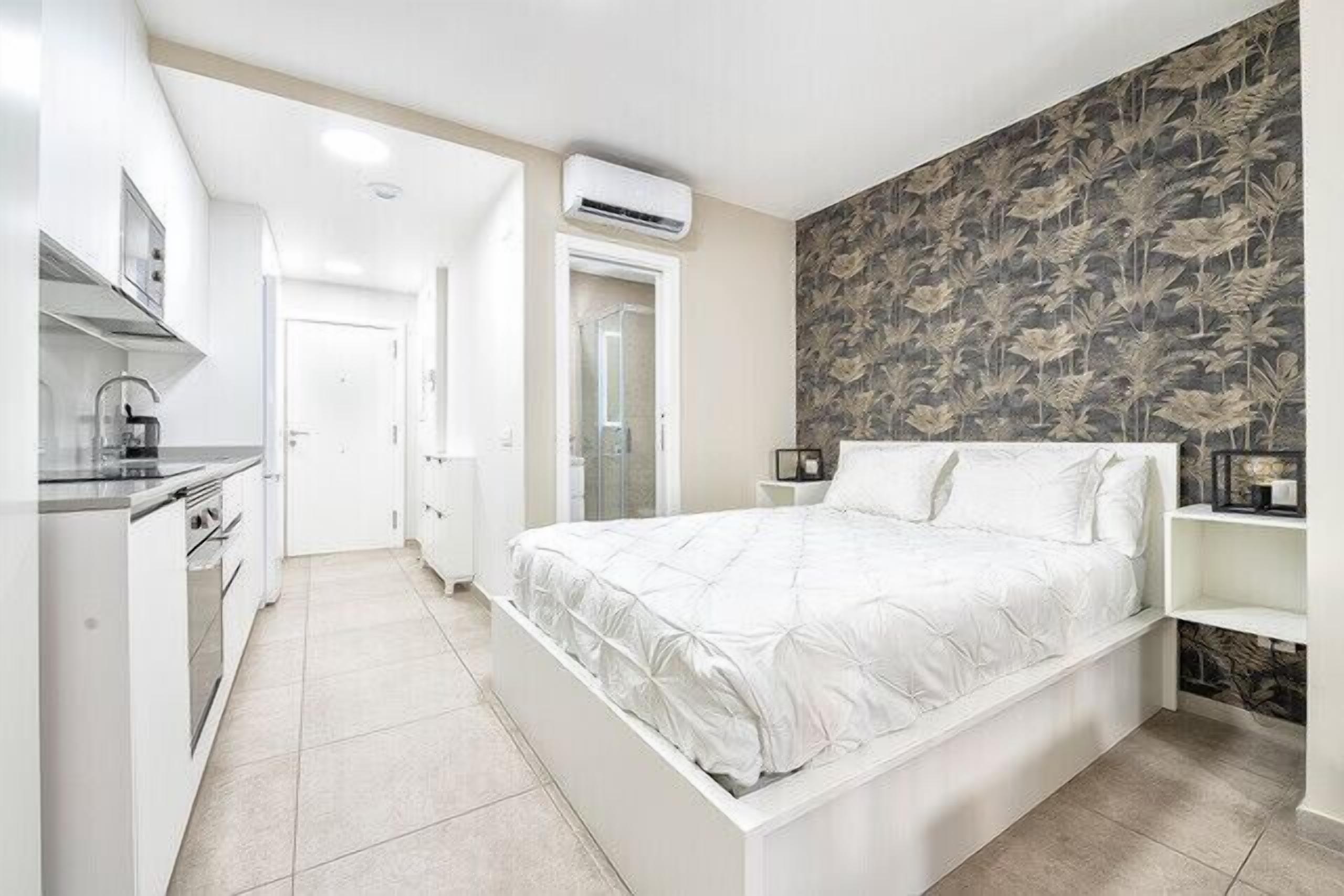 Un dormitorio con cama blanca, una mesita de noche pequeña, un armario y un baño con ducha e inodoro.
