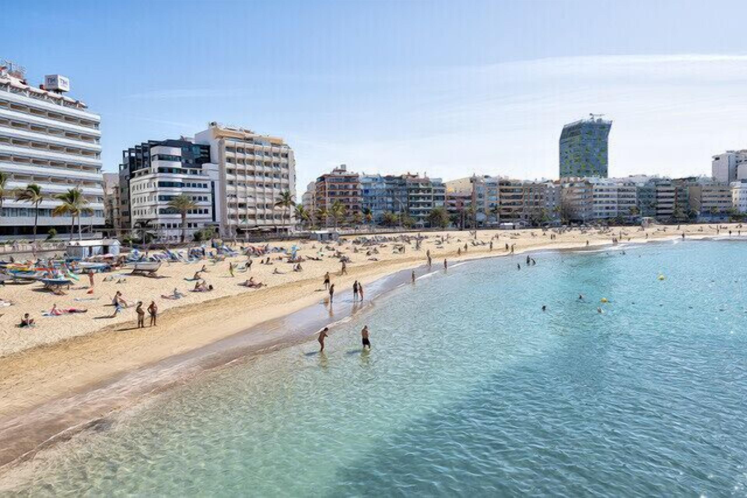 A beach with clear water, a sandy shore, and buildings in the background.