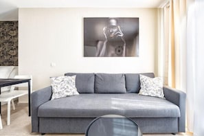 Ein graues Sofa mit gemusterten Kissen, ein gerahmtes Kunstwerk darüber und ein Esstisch mit Stühlen links davon.