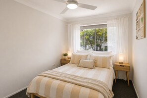 A bedroom with a bed, a ceiling fan, a window with curtains, and bedside lamps. - The Burleigh Hideaway (Gold Coast)