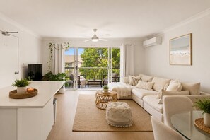 Uma sala de estar moderna com sofá, mesa de centro e mesa de jantar de vidro. Há um ventilador de teto, uma televisão e uma janela com cortinas.