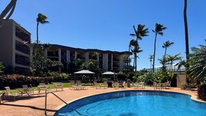 Una piscina di un resort con lettini e ombrelloni, circondata da palme e un edificio a più piani.