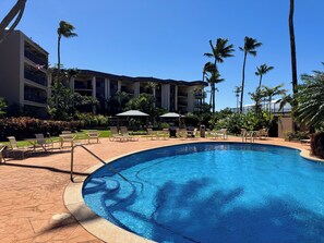 Ein Hotelpool mit Liegestühlen und Sonnenschirmen, umgeben von Palmen und einem mehrstöckigen Gebäude.