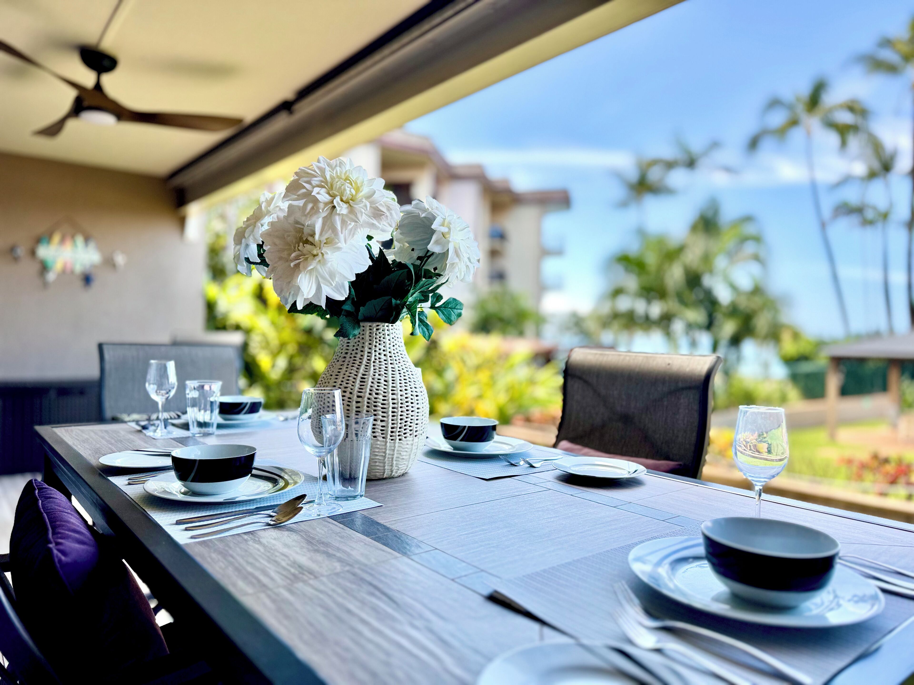 A dining table set for two with a vase of flowers, a ceiling fan, and a view of palm trees.