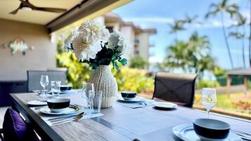 A dining table set for two with a vase of flowers, a ceiling fan, and a view of palm trees.