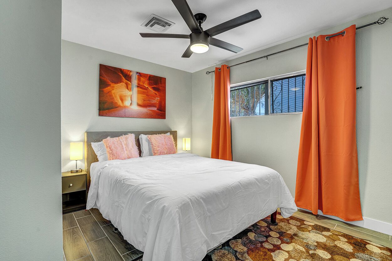 Un dormitorio con cama, ventilador de techo, una obra de arte en la pared y una ventana con cortinas.