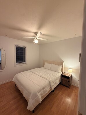 A bedroom with a bed, a nightstand, a ceiling fan, a window with blinds, and a wall-mounted mirror.