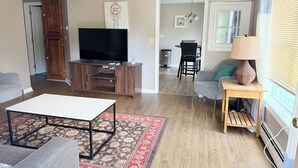 A living room with a flat-screen TV, a sofa, a coffee table, and a dining area in the background. - Nestled in the trees. (Saco)