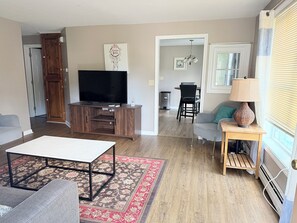 A living room with a flat-screen TV, a sofa, a coffee table, and a dining area in the background. - Nestled in the trees. (Saco)