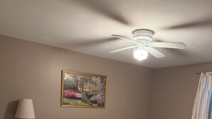 A bedroom with a bed, a nightstand, a lamp, a ceiling fan, a window with curtains, and a wall-mounted painting. - Nestled in the trees. (Saco)