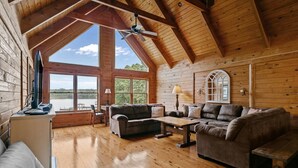 Interior de una cabaña de madera con techo alto, ventanas amplias, chimenea y una zona de estar acogedora.
