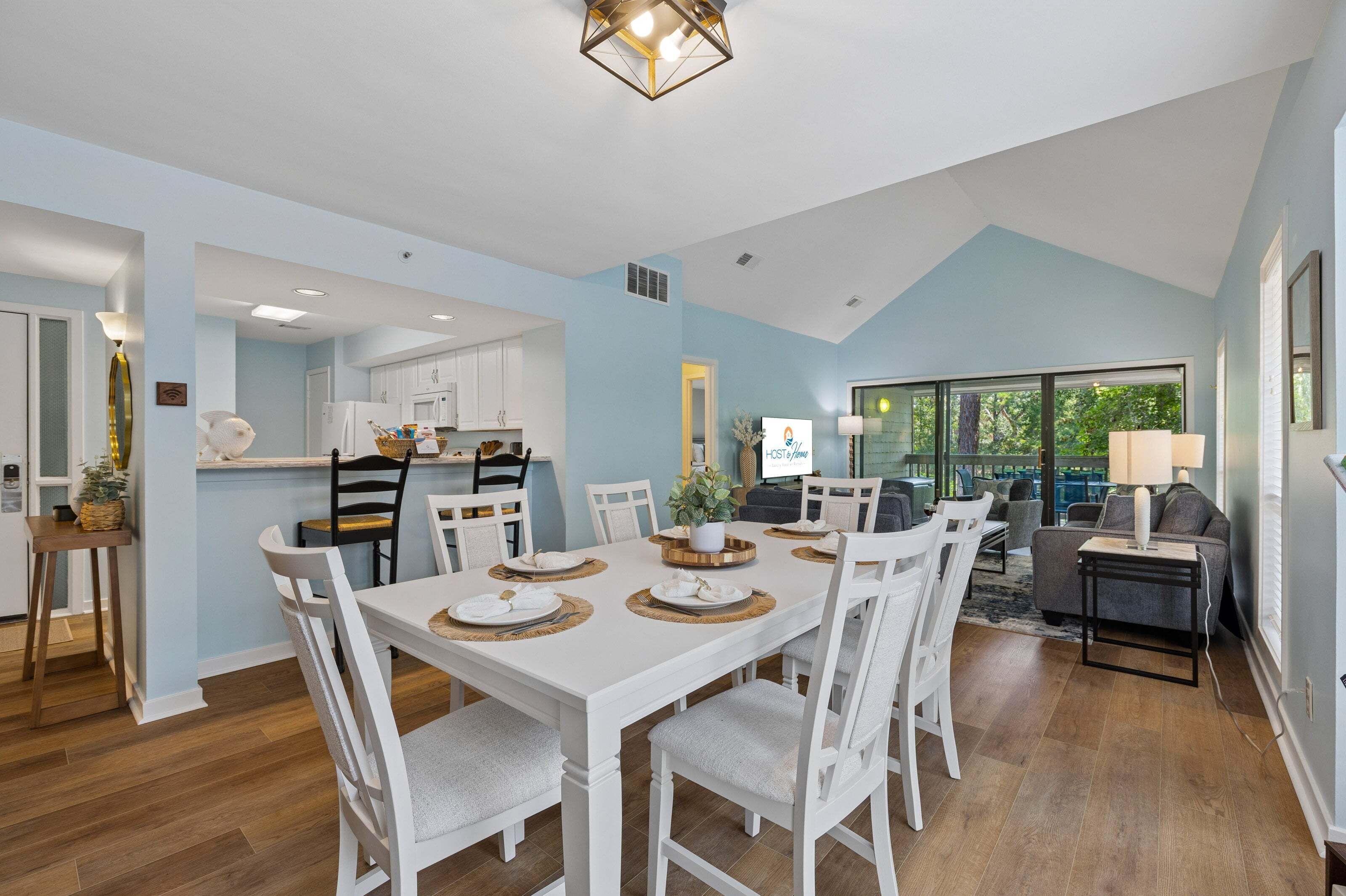 A modern dining area with a white table set for four, a kitchen in the background, and a view of the outdoors.