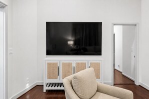Un salon avec un grand téléviseur à écran plat fixé à un meuble blanc, un fauteuil beige et un plancher en bois.