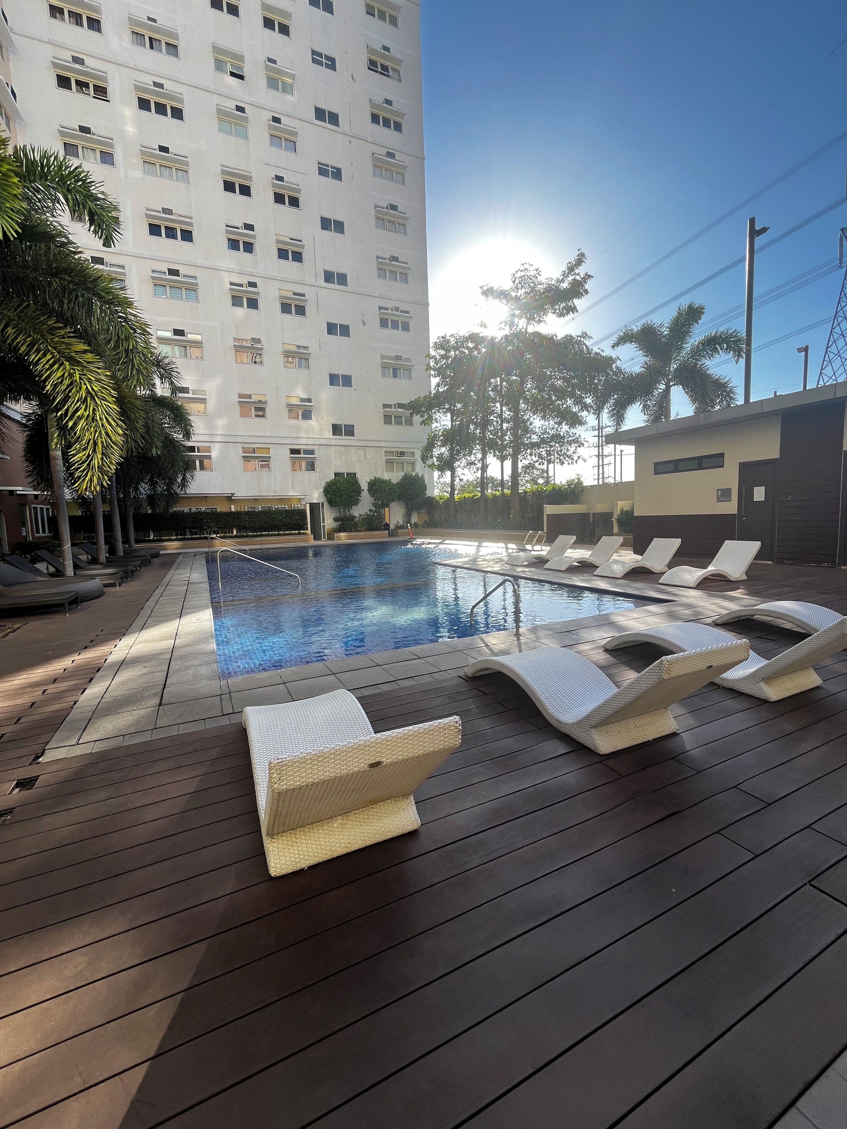 Una piscina con sillones de descanso, una terraza de madera y un edificio alto al fondo.