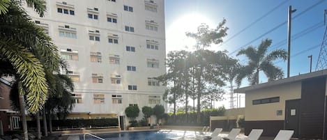Una piscina con sillones de descanso, una terraza de madera y un edificio alto al fondo.