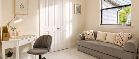 Una habitación con escritorio, silla y sofá, una puerta blanca y una ventana con persianas.
