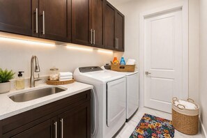 A compact kitchen with dark wooden cabinets, a stainless steel sink, a built-in washing machine, and a small laundry basket. - NEW! Canal Escape, Pool, Kayaks & Outdoor Kitchen (Cape Coral)
