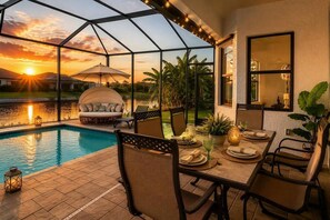A poolside dining area with a glass enclosure, a cushioned seating area, and a view of the sunset over a body of water. - NEW! Canal Escape, Pool, Kayaks & Outdoor Kitchen (Cape Coral)