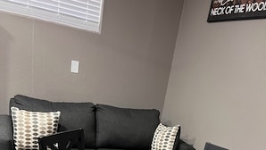 Una sala de estar moderna con una mesa de centro de vidrio, dos sillas de madera, un sofá gris oscuro y cojines estampados.