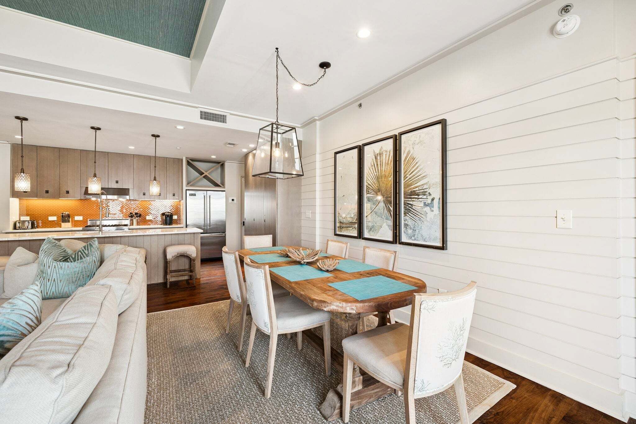 A modern kitchen with pendant lights, a dining area with a wooden table and chairs, and a living room with a sofa and cushions.