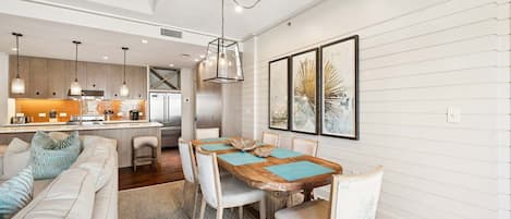 A modern kitchen with pendant lights, a dining area with a wooden table and chairs, and a living room with a sofa and cushions.
