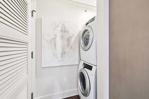 A laundry room with a wall-mounted washer and dryer, a white door with horizontal slats, and a large abstract painting on the wall. - 6 Barrett Place | Elevated Luxury in the Heart of Rosemary Beach (Rosemary Beach)