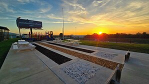 Ein Spielplatz mit Rutschen und einem Picknickbereich, ein Hinweisschild und ein Sonnenuntergang im Hintergrund.