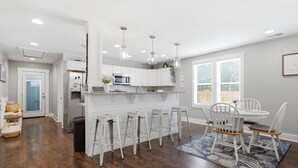 Una cocina moderna con armarios blancos, una isla central con taburetes de bar, iluminación colgante y un comedor con sillas y mesa blancos.
