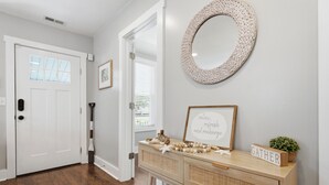 Un pasillo con piso de madera, una puerta blanca, un espejo, una consola decorativa y un letrero que dice 'GATHER'.