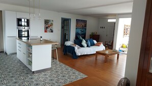 Eine moderne Küche mit einer zentralen Kochinsel, ein Essbereich mit Holztisch, eine Couch mit blauen Kissen und ein gemustertes Teppich.