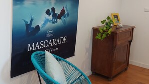 Ein Zimmer mit einem 'MascaraDE'-Poster an der Wand, einem blauen Stuhl, einem Holzschrank und einer kleinen Pflanze.
